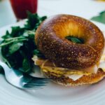 a bagel sandwich with eggs and cheese with spinach and a fork on a white plate | breakfast lone tree